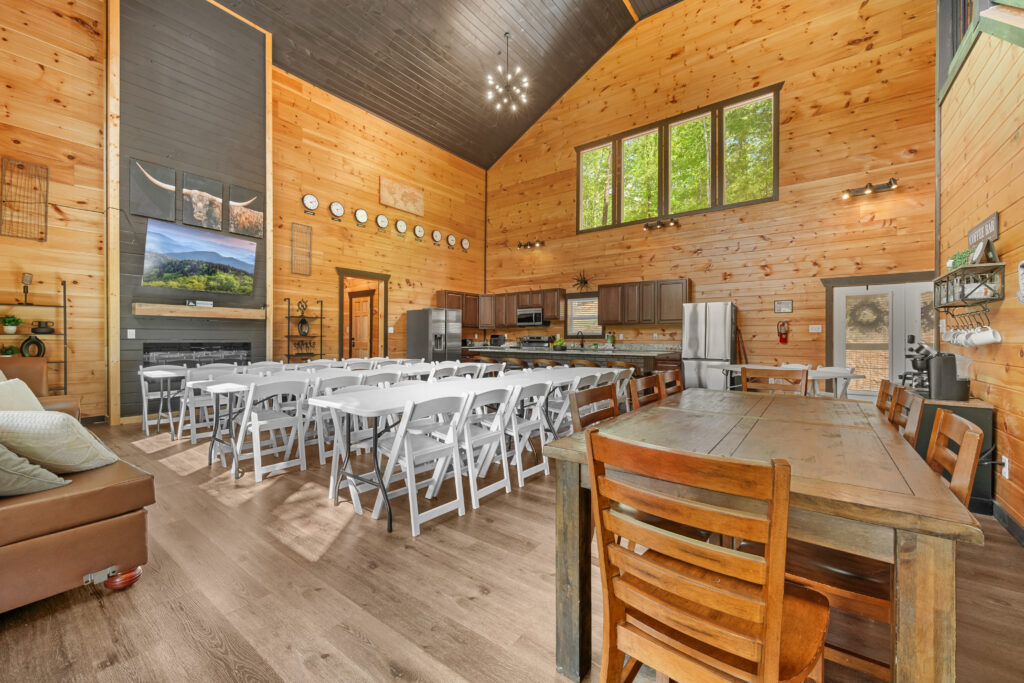 Spacious lodge dining area with wooden walls, large windows, long tables with white chairs, a kitchen in the back, and a rustic wooden dining table in the foreground—perfect for gatherings after a day exploring Smoky Mountain Cabins or enjoying the warmth of nearby Cabin Communities.