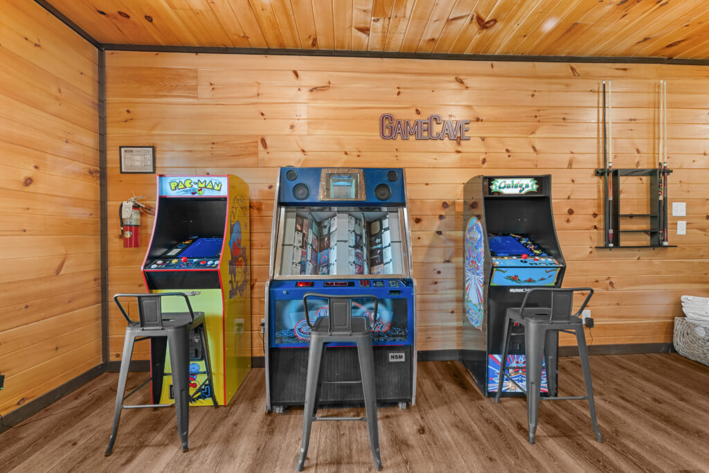 Three arcade machines—Pac-Man, a racing game, and Galaga—are lined up against a wooden wall in a cozy cabin game room. Three metal stools are placed in front of the machines, and pool cues hang on the right, creating the perfect spot for fun and relaxation in the heart of Smoky Mountain communities.